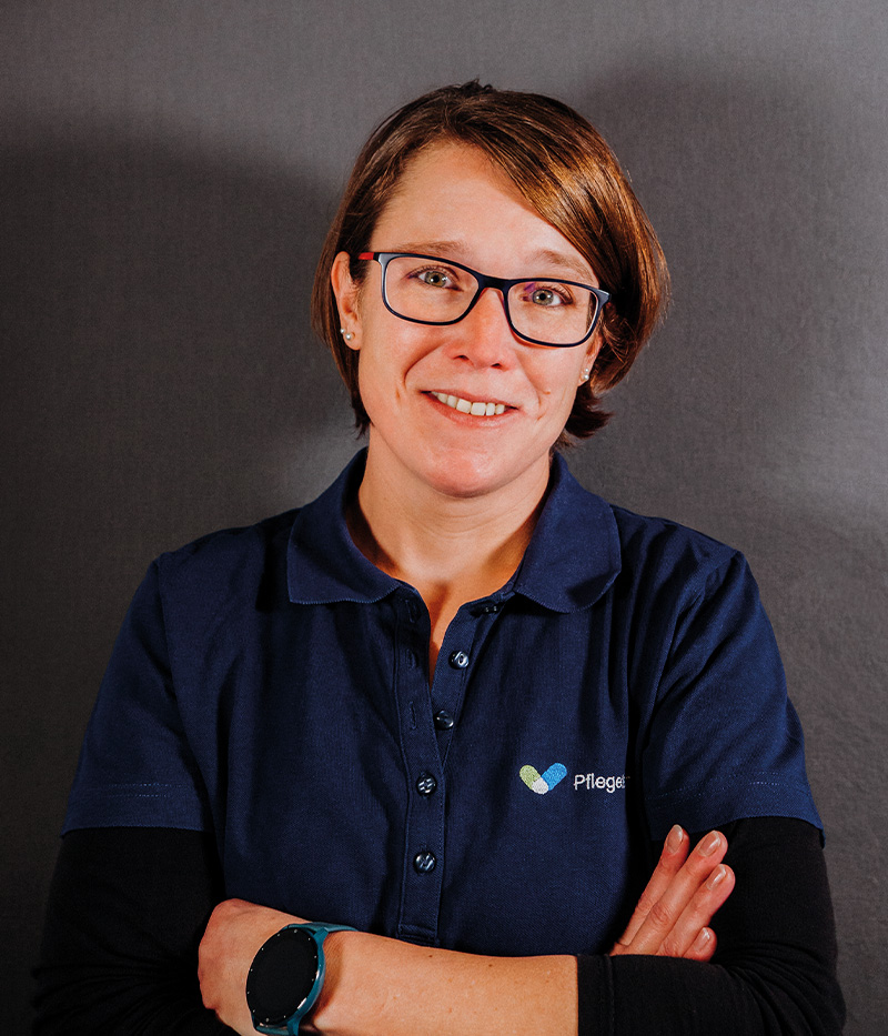 Stefanie Halm, Betreuungs- und Hauswirtschaftskraft beim Pflegeteam Drensteinfurt, lächelt freundlich in die Kamera. Sie trägt ein dunkelblaues Poloshirt mit Logo und steht mit verschränkten Armen vor grauem Hintergrund.