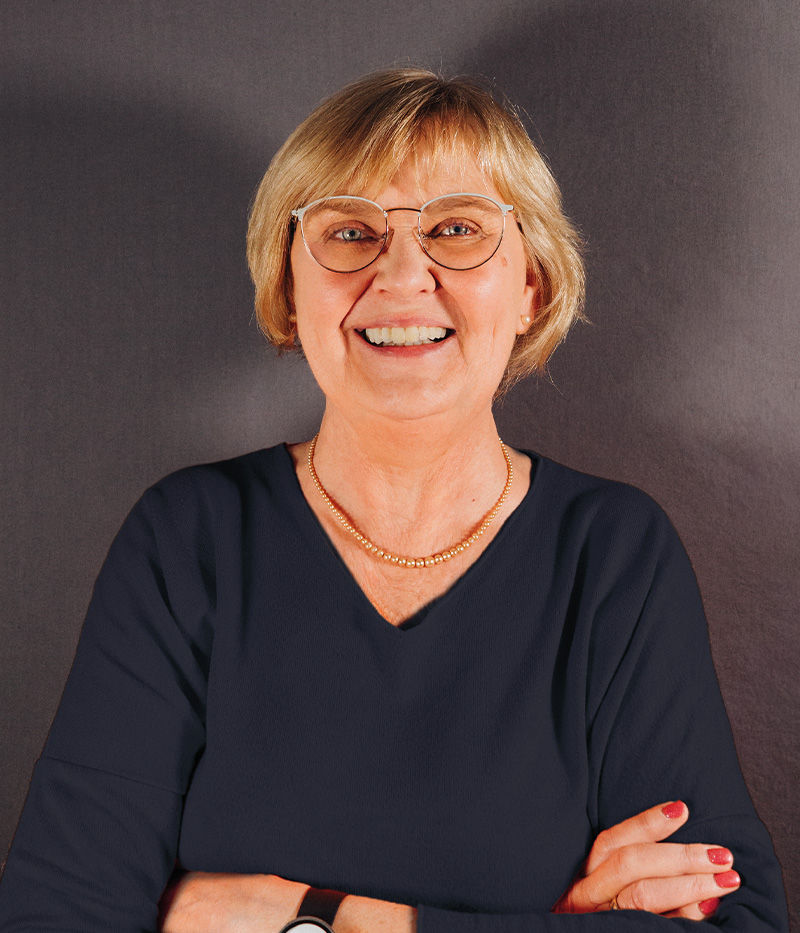 Elvira Niggemann, Krankenschwester und QM-Beauftragte beim Pflegeteam Drensteinfurt, lächelt freundlich in die Kamera. Sie trägt eine runde Brille, ein dunkelblaues Oberteil und eine goldene Halskette vor grauem Hintergrund.