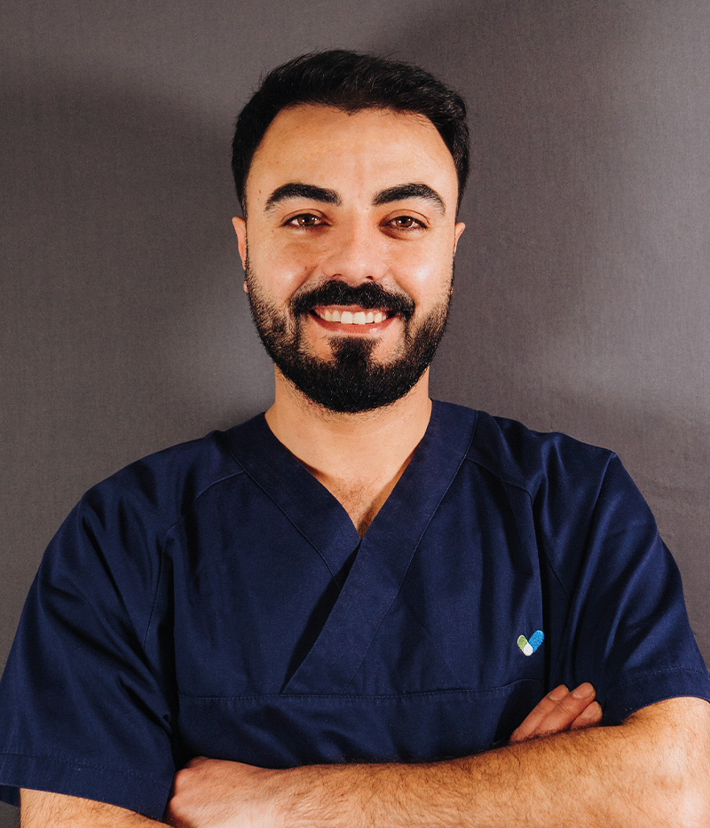 Nihat Tamo, Pflegefachmann beim Pflegeteam Drensteinfurt, lächelt freundlich in die Kamera. Er trägt dunkelblaue Pflegekleidung und steht mit verschränkten Armen vor grauem Hintergrund.